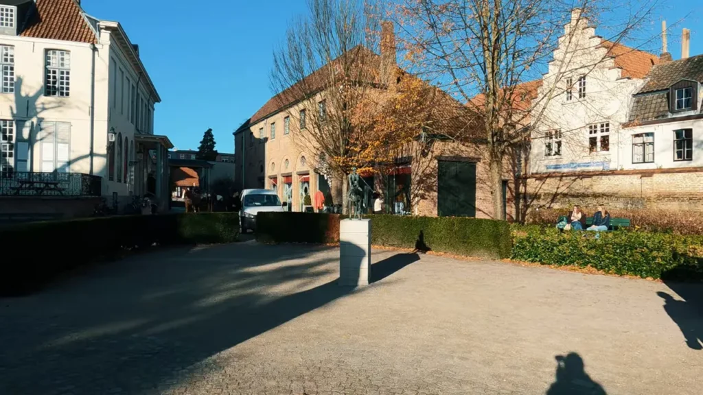 Praça em Bruges com escultura sobre pedestal, árvores e edifícios históricos ao fundo
