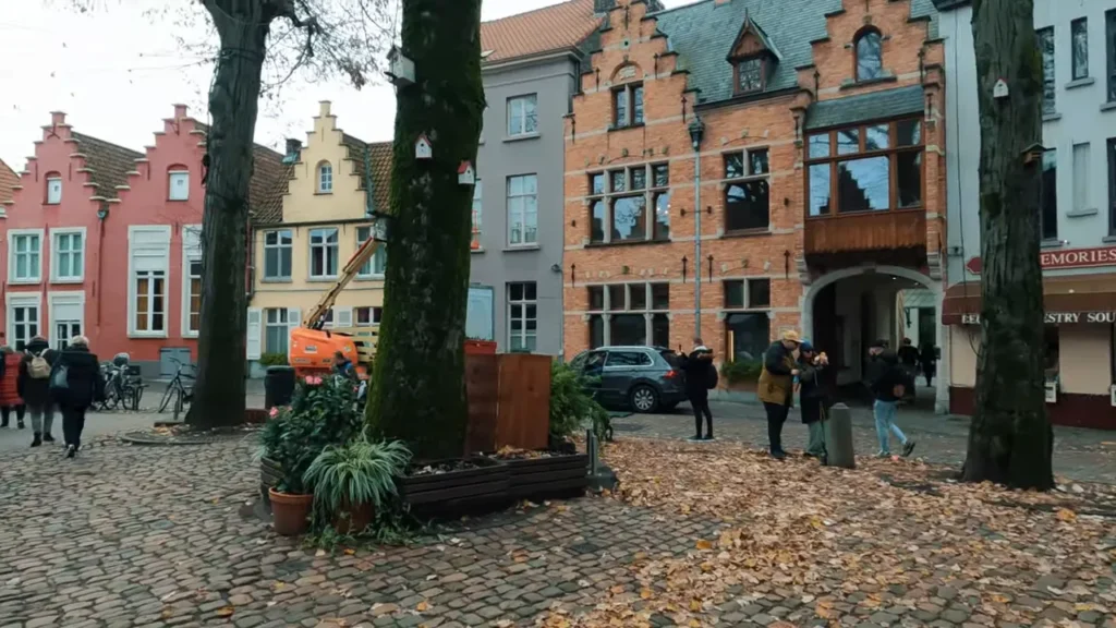 Praça histórica de Bruges com árvores, fachadas de tijolos e pessoas caminhando