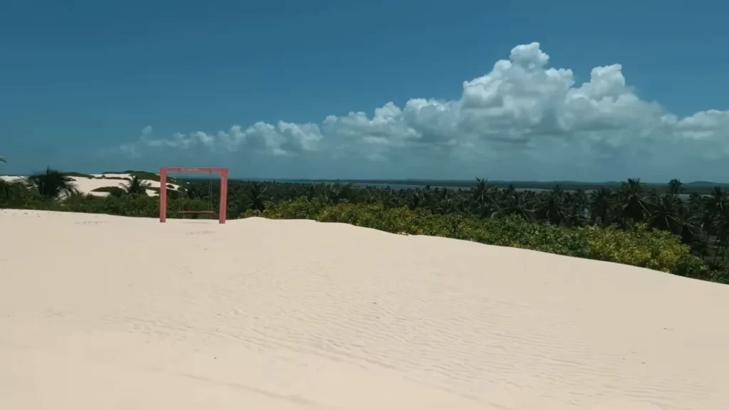 Duna ampla com balanço vermelho em primeiro plano e mata costeira ao fundo sob céu azul.