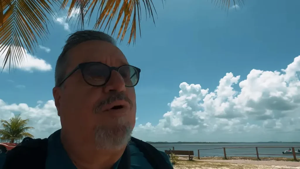 Vista da orla com lagoa ao fundo, banco, coqueiro e céu azul com nuvens — cenário do passeio