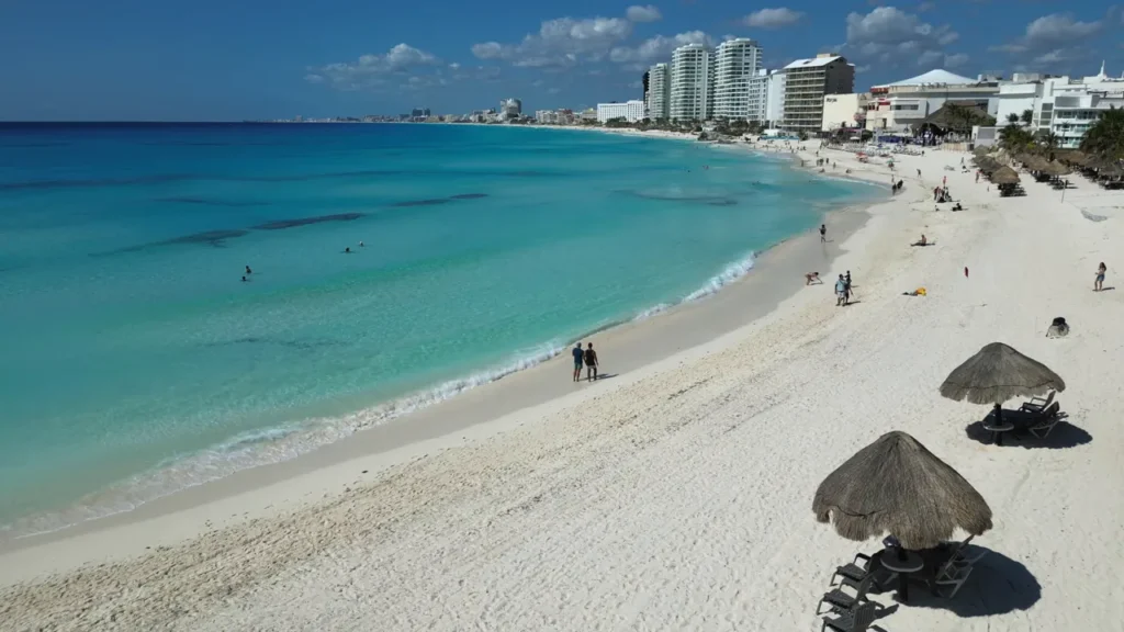 Praia de areia branca com palapas e mar azul claro, mostrando hotelaria ao fundo